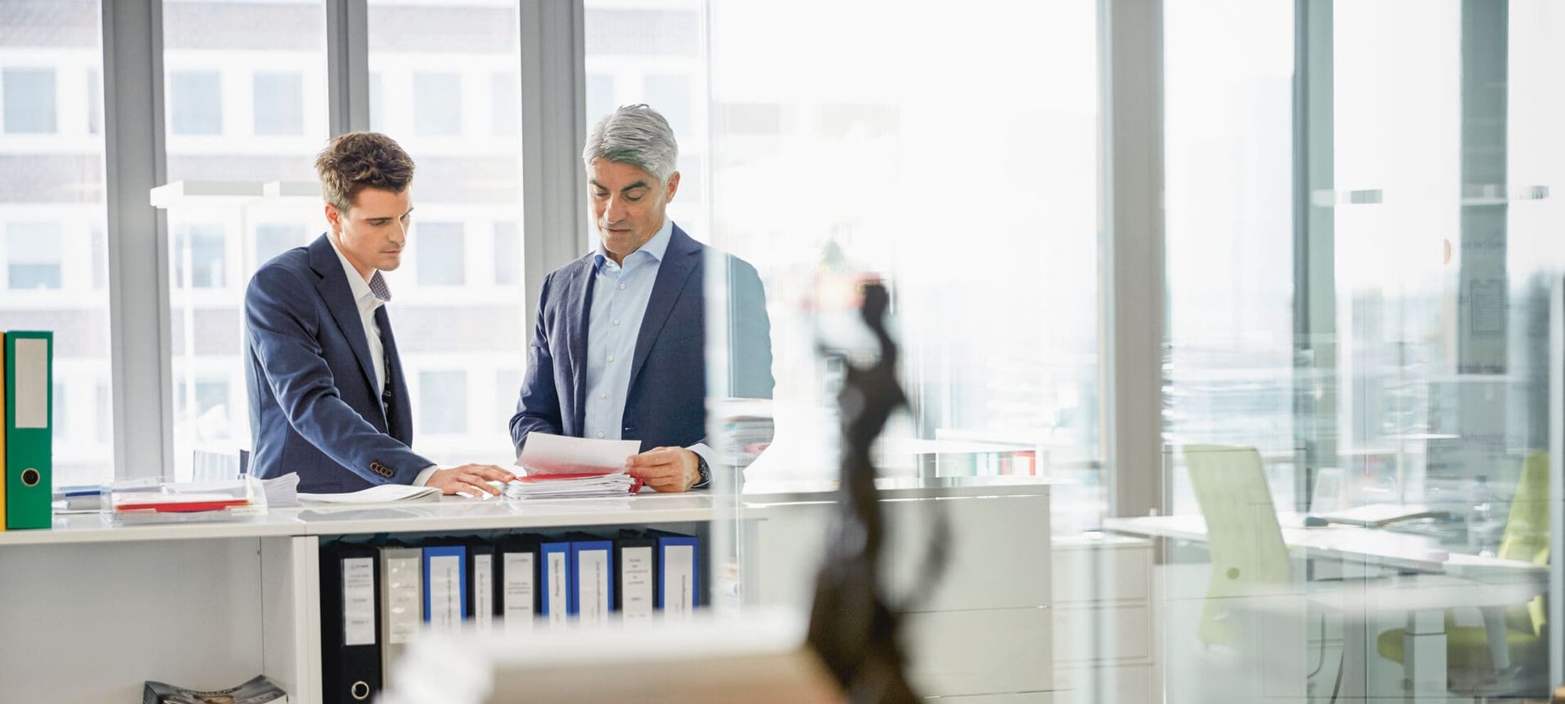 Deux hommes en costume discutent de questions juridiques. Au premier plan, on voit floue une figure de Justitia. 