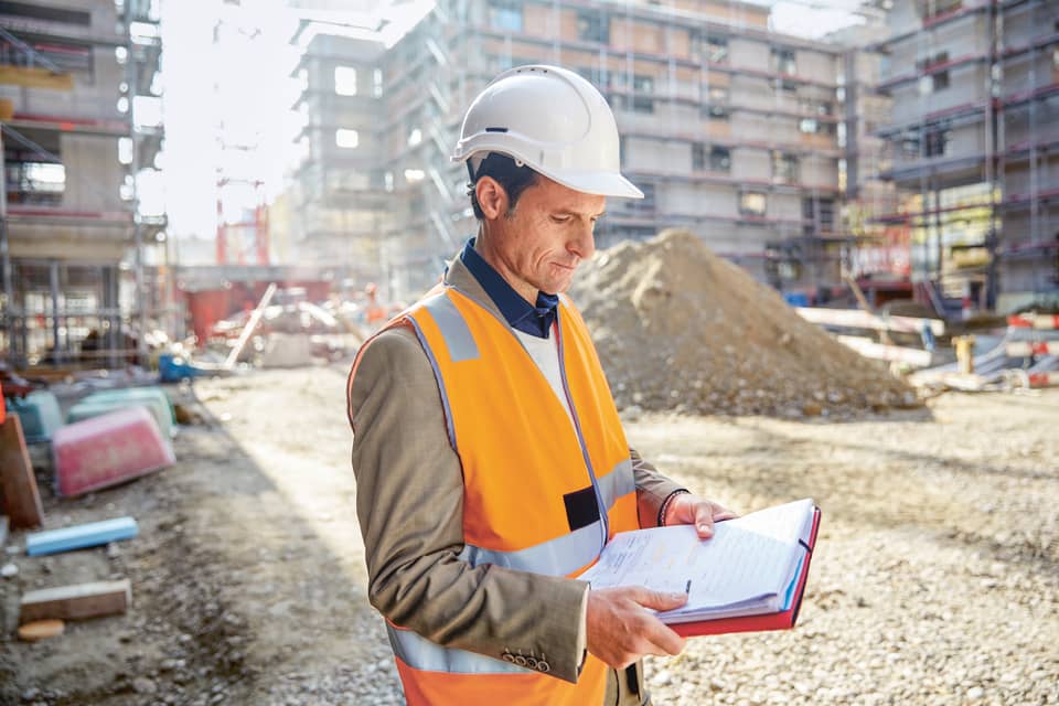 Mann mit Sicherheitshelm und Weste überprüft Dokumente auf einer Baustelle